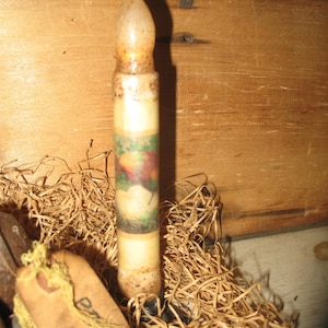 May include: A vintage-style candle with a painted fruit design. The candle has a cream-colored body with a detailed image of apples and pears. It is surrounded by straw and set against a wooden background, evoking a rustic aesthetic.