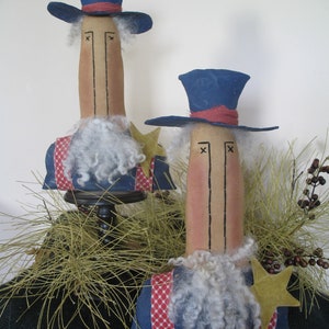 May include: Two handmade patriotic dolls with blue top hats, red, white, and blue clothing, and white beards. The dolls are standing on a base with a brown surface and green foliage.