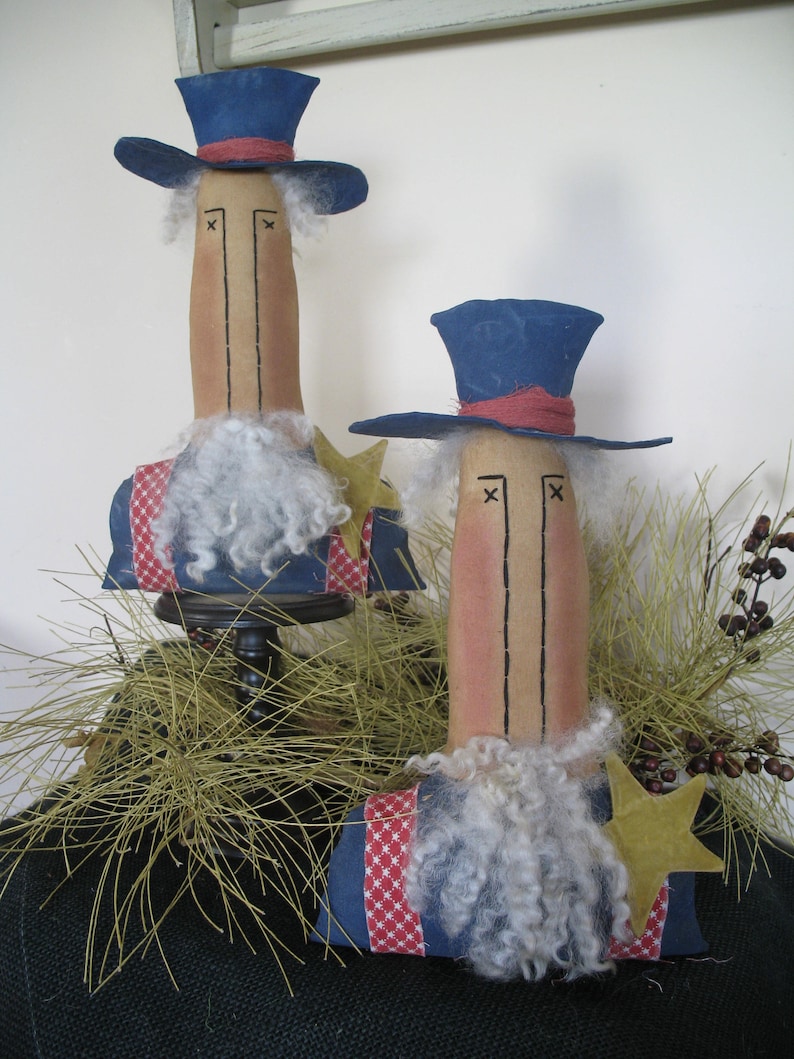 May include: Two handmade patriotic dolls with blue top hats, red, white, and blue clothing, and white beards. The dolls are standing on a base with a brown surface and green foliage.