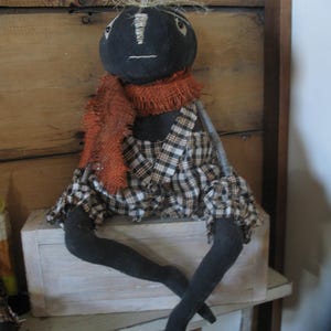 K&ouml;nnte beinhalten: Eine schwarze Stoffpuppe mit einem karierten Rock, einem orangefarbenen Schal und &Auml;sten als Armen. Die Puppe sitzt auf einer wei&szlig;en Holzkiste.