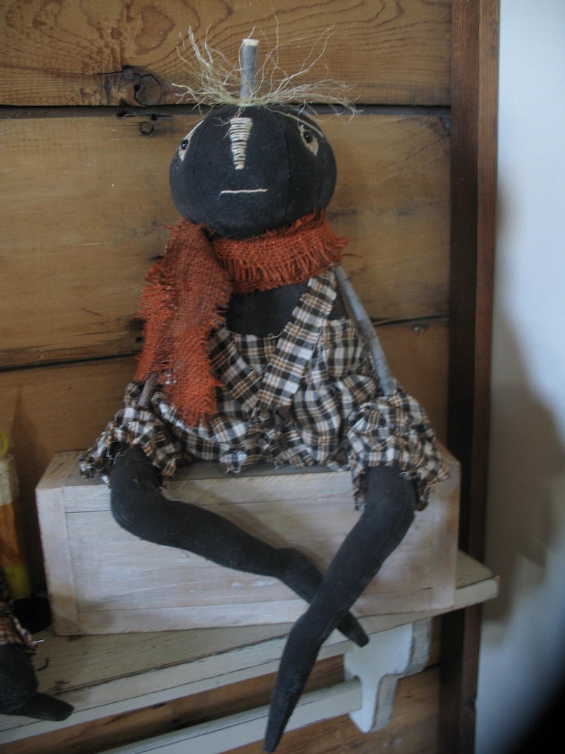 K&ouml;nnte beinhalten: Eine schwarze Stoffpuppe mit einem karierten Rock, einem orangefarbenen Schal und &Auml;sten als Armen. Die Puppe sitzt auf einer wei&szlig;en Holzkiste.