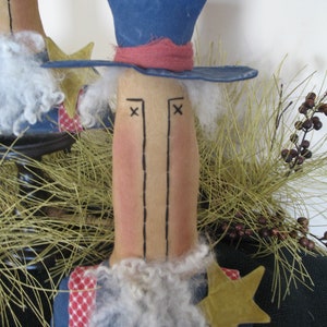 May include: A handmade rag doll with a blue hat, red and white striped fabric, and a white beard. The doll has a brown face with black eyes and a stitched-on mouth. The doll is standing in front of a brown and green background.