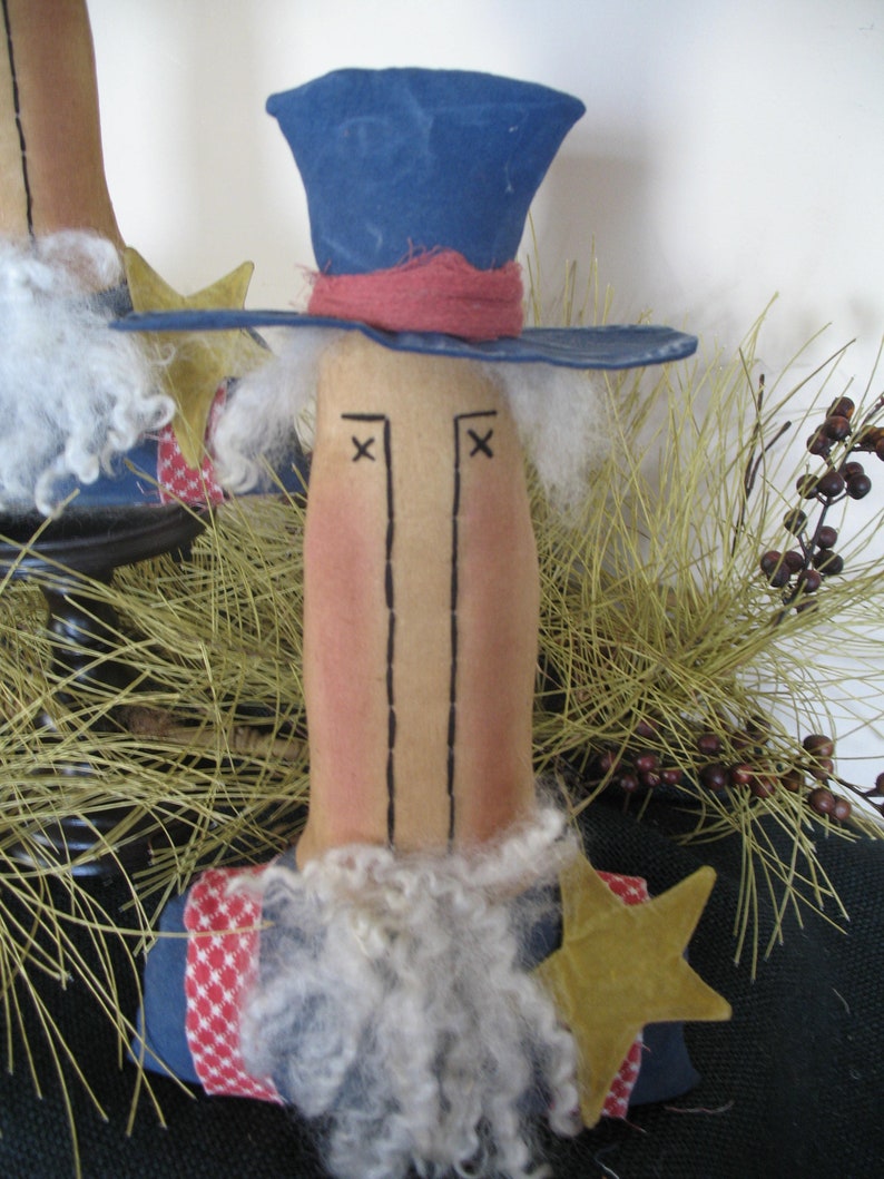May include: A handmade rag doll with a blue hat, red and white striped fabric, and a white beard. The doll has a brown face with black eyes and a stitched-on mouth. The doll is standing in front of a brown and green background.