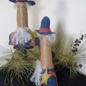 May include: Two handmade fabric dolls dressed as Uncle Sam. The dolls are standing on a bed of dried grass. The taller doll is wearing a blue top hat, a blue jacket, and a white beard. The shorter doll is wearing a blue hat, a blue jacket, and a red, white, and blue star.