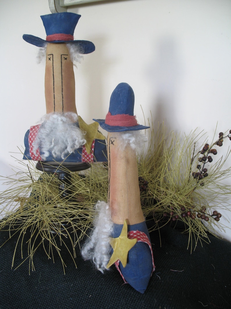 May include: Two handmade fabric dolls dressed as Uncle Sam. The dolls are standing on a bed of dried grass. The taller doll is wearing a blue top hat, a blue jacket, and a white beard. The shorter doll is wearing a blue hat, a blue jacket, and a red, white, and blue star.