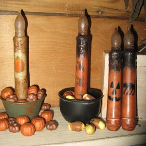 May include: A collection of Halloween-themed candles and decorations. The candles are orange and brown, with jack-o'-lantern and spider designs. Small orange and brown pumpkins and corn kernels are scattered around the candles.
