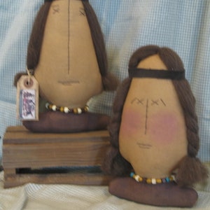 May include: Two handmade rag dolls with brown hair, brown eyes, and beaded necklaces. The dolls are wearing brown headbands and are sitting on a wooden crate.