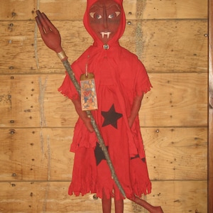 May include: A handmade red devil doll with a horned hood, holding a staff with a hand-shaped top. The doll has a black star on its dress and a tag hanging from its neck. The background is a wooden wall.