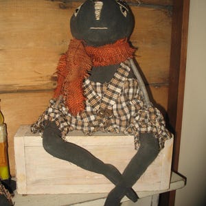 K&ouml;nnte beinhalten: Eine schwarze Stoffpuppe mit einem karierten Kleid und einem orangefarbenen Schal sitzt auf einer wei&szlig;en Holzkiste. Die Puppe hat einen Zweig als Nase und stroh&auml;hnliches Haar.