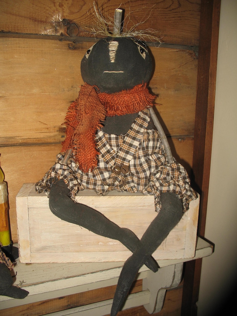 K&ouml;nnte beinhalten: Eine schwarze Stoffpuppe mit einem karierten Kleid und einem orangefarbenen Schal sitzt auf einer wei&szlig;en Holzkiste. Die Puppe hat einen Zweig als Nase und stroh&auml;hnliches Haar.