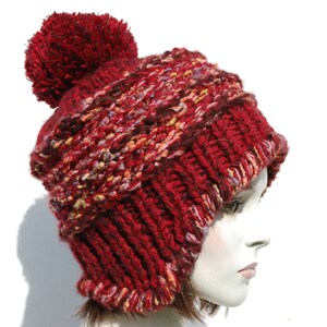 May include: A red knitted beanie with a fluffy pom pom on top. The beanie has a multi-coloured pattern with shades of red, pink, and white.