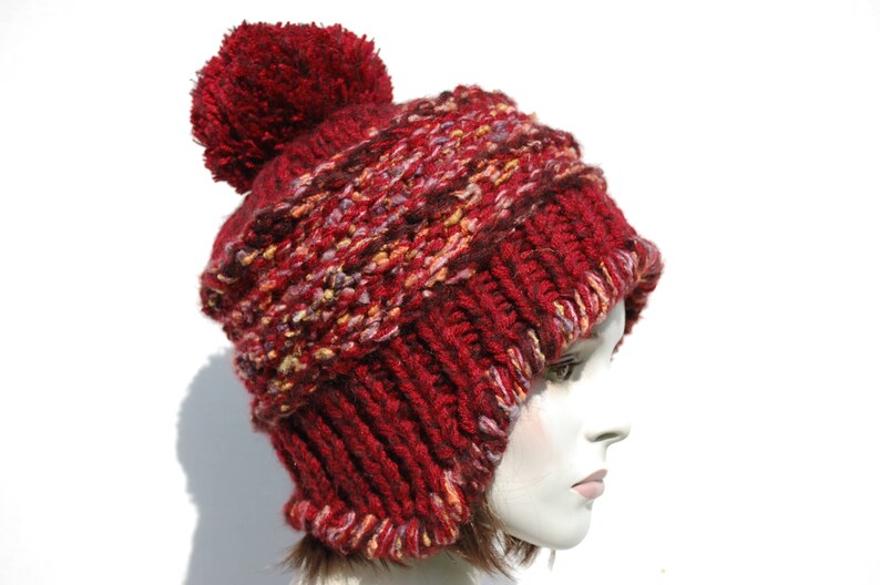 May include: A red knitted beanie with a fluffy pom pom on top. The beanie has a multi-coloured pattern with shades of red, pink, and white.
