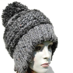 May include: A grey knitted hat with a fluffy pom pom on top. The hat has a long flap that can be worn down over the ears or up on top of the head.