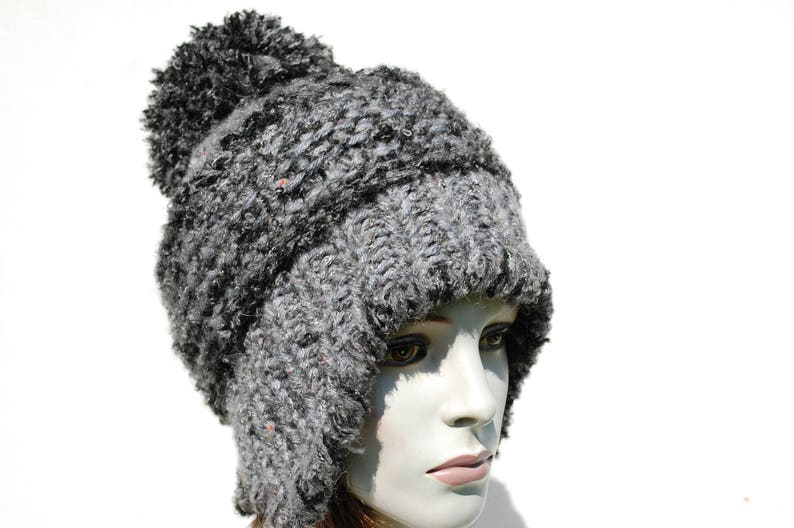 May include: A grey knitted hat with a fluffy pom pom on top. The hat has a long flap that can be worn down over the ears or up on top of the head.