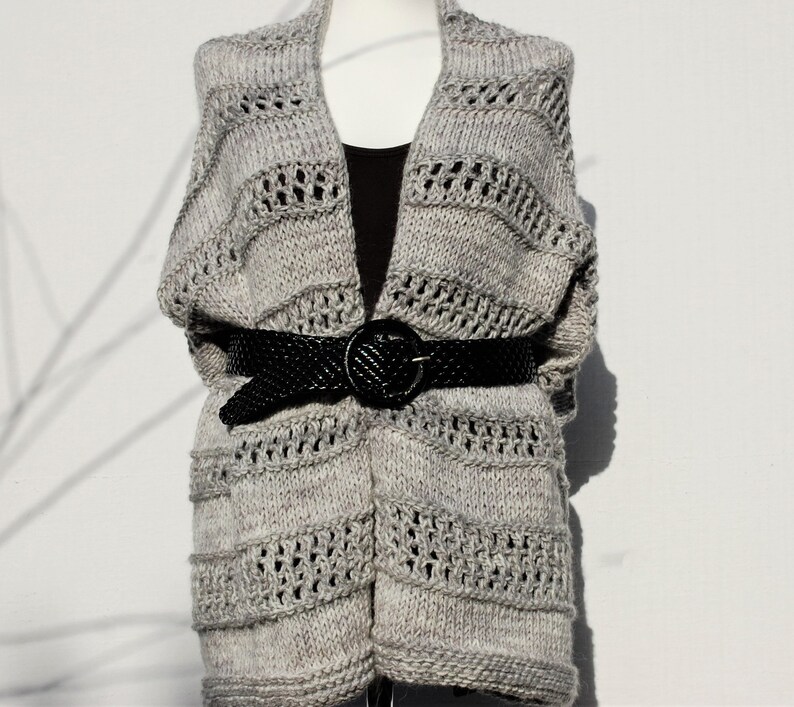 May include: A gray knitted cardigan with a open front and a black belt. The cardigan has a textured knit pattern with a lace-like design.