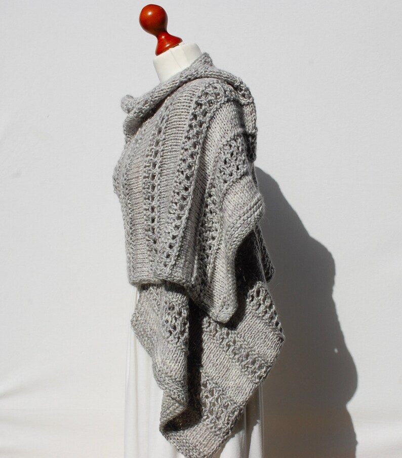 May include: A gray knitted poncho with a cowl neck and a lacy pattern. The poncho is draped over a mannequin.