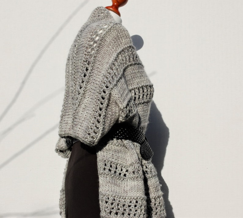 May include: A gray knitted cardigan with a delicate openwork pattern. The cardigan is draped over a mannequin and has a black belt.