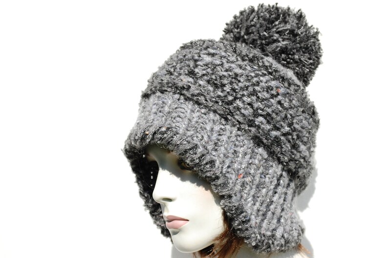 May include: A grey and black fuzzy knit hat with a pom pom on top. The hat has a long flap that can be pulled down to cover the ears.