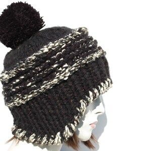 May include: A black and white knitted beanie with a large black pom pom on top. The beanie has a thick, textured knit and a folded brim.