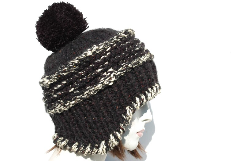May include: A black and white knitted beanie with a large black pom pom on top. The beanie has a thick, textured knit and a folded brim.