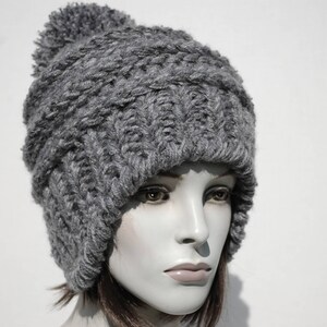 May include: A grey knitted beanie with a fluffy pom pom on top. The beanie has a cable knit pattern and is folded down at the brim.