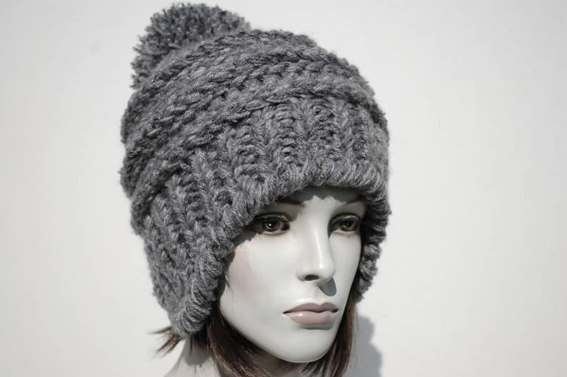 May include: A grey knitted beanie with a fluffy pom pom on top. The beanie has a cable knit pattern and is folded down at the brim.