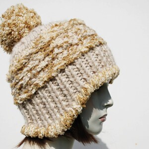 May include: A beige and brown knitted beanie with a fluffy pom pom on top. The beanie has a cable knit pattern and a fuzzy texture.