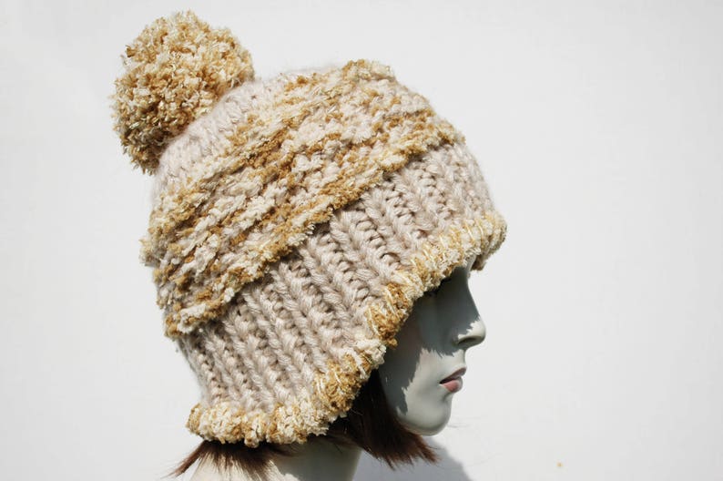 May include: A beige and brown knitted beanie with a fluffy pom pom on top. The beanie has a cable knit pattern and a fuzzy texture.