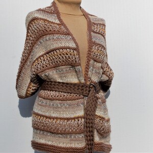 May include: A brown, white, and beige striped knitted cardigan with a brown braided belt. The cardigan has a V-neck and long sleeves.