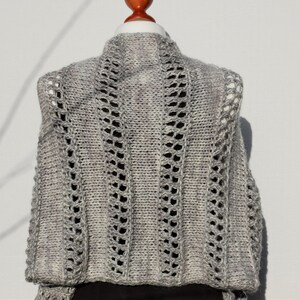 May include: A gray knitted vest with a lacy pattern. The vest has a high neckline and a loose fit.
