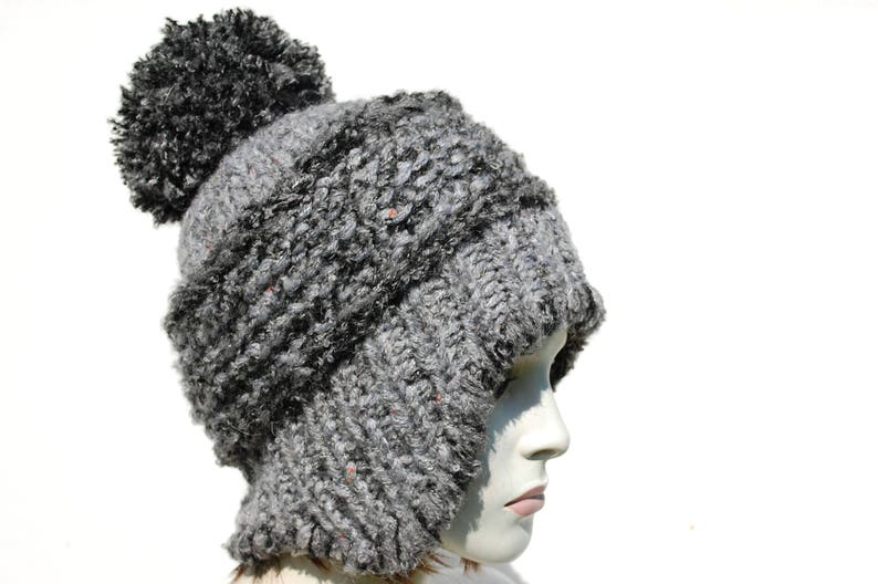May include: A grey knitted hat with a large pom-pom on top. The hat has a fold-down ear flap design.