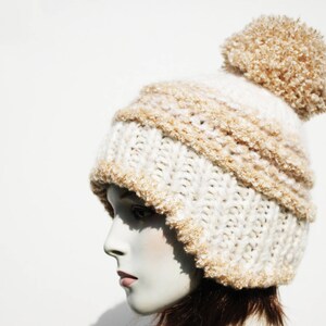 May include: A white and beige knitted beanie with a fluffy pom pom on top. The beanie is made of a soft, fuzzy yarn and has a slouchy fit.