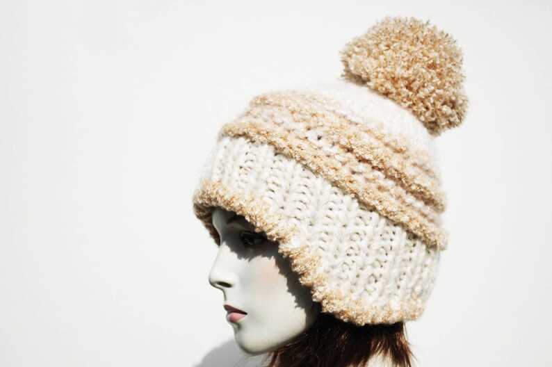 May include: A white and beige knitted beanie with a fluffy pom pom on top. The beanie is made of a soft, fuzzy yarn and has a slouchy fit.