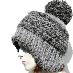 May include: A grey and black knitted beanie with a fluffy pom pom on top. The beanie has a fold-down flap that covers the ears.
