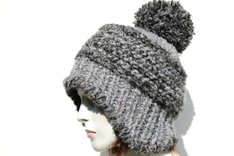 May include: A grey and black knitted beanie with a fluffy pom pom on top. The beanie has a fold-down flap that covers the ears.