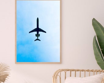 Puede incluir: Una impresión enmarcada que muestra un avión de pasajeros desde abajo contra un cielo azul. La obra de arte está en un marco de madera clara. La imagen está en una habitación con una silla de mimbre, un cojín blanco y una alfombra de tonos neutros.