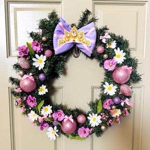 May include: A decorative wreath with a green base, adorned with pink and white flowers, glittery pink ornaments, and a purple bow with a gold crown. The wreath is designed for door decor.