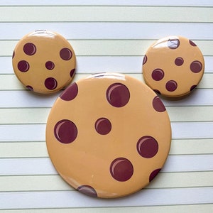 May include: Three round, brown and tan cookie-shaped buttons with a chocolate chip design. The largest button is in the center and the two smaller buttons are on either side.