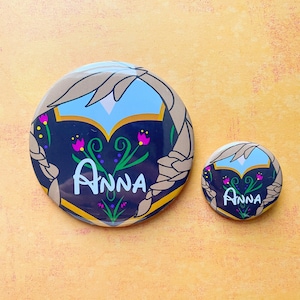 May include: Two round buttons with a black background and a white outline. The larger button has a floral design with pink flowers and the word "ANNA" in white. The smaller button has a similar design with the word "ANNA" in white.