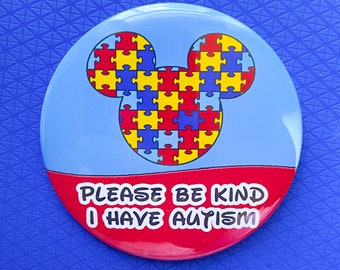 May include: A round button with a light blue background and a red bottom. The button features a Mickey Mouse head made of puzzle pieces in red, yellow, and blue. Text on the red section reads "PLEASE BE KIND I HAVE AUTISM."