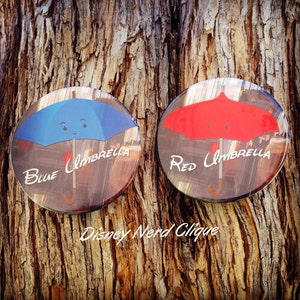 May include: Two round buttons with images of blue and red umbrellas. The text "Blue Umbrella" and "Red Umbrella" is written on each button. The text "Disney Nerd Clique" is written below the buttons.