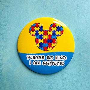May include: A yellow and blue button with a Mickey Mouse head made of puzzle pieces. The text on the button reads "Please be kind I am autistic."