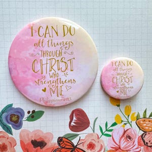 May include: Two pink and white watercolor-style round buttons with gold lettering. The larger button reads "I can do all things through Christ who strengthens me Philippians 4:13". The smaller button reads "I can do all things through Christ who strengthens me Philippians 4:13".
