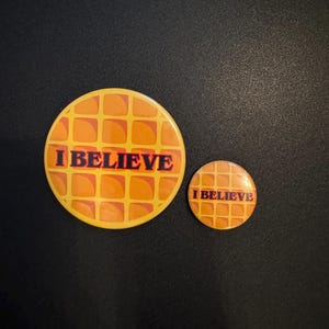 May include: Two round buttons with a waffle design in shades of orange and yellow. The text "I BELIEVE" is printed in black on each button. One button is larger than the other.