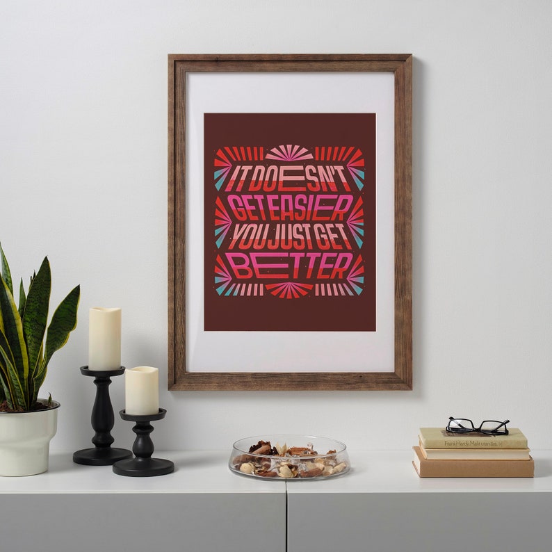 May include: A framed print with a motivational quote in a colorful retro style. The quote reads "It doesn't get easier, you just get better". The print is framed in a brown wood frame and is displayed on a shelf with candles and a bowl of decorative items.