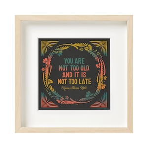 May include: A framed print with a black background and a colourful floral border. The text in the print reads "You are not too old and it is not too late." The quote is attributed to Rainer Maria Rilke.