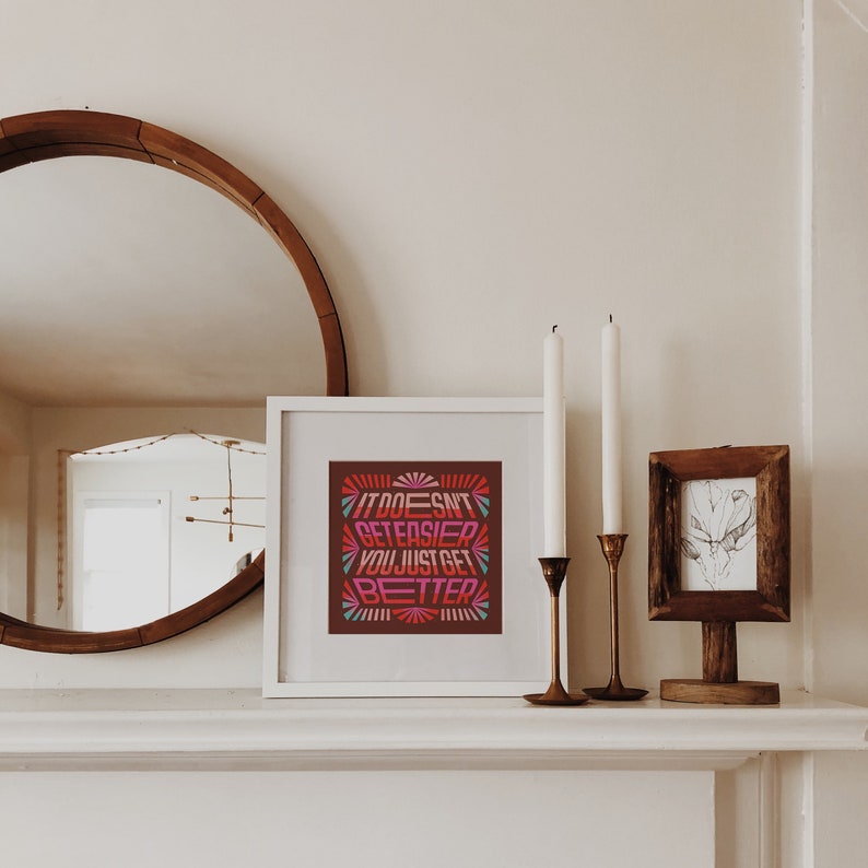 May include: A white framed print with a motivational quote in a colorful retro font. The quote reads "It Doesn't Get Easier You Just Get Better". The print is displayed on a white mantel with two candlesticks and a small framed print.
