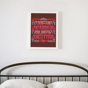 May include: A framed print with a motivational quote in a retro style font. The quote reads "It doesn't get easier, you just get better." The background is a dark brown color with a colorful design.