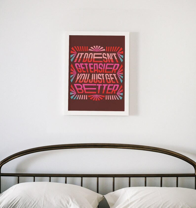 May include: A framed print with a motivational quote in a retro style font. The quote reads "It doesn't get easier, you just get better." The background is a dark brown color with a colorful design.