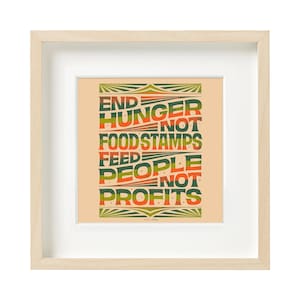 May include: Framed art print with the text "END HUNGER NOT FOOD STAMPS FEED PEOPLE NOT PROFITS" in green, orange, and red. The print is set against a peach background, framed in a light wood frame with a white mat.